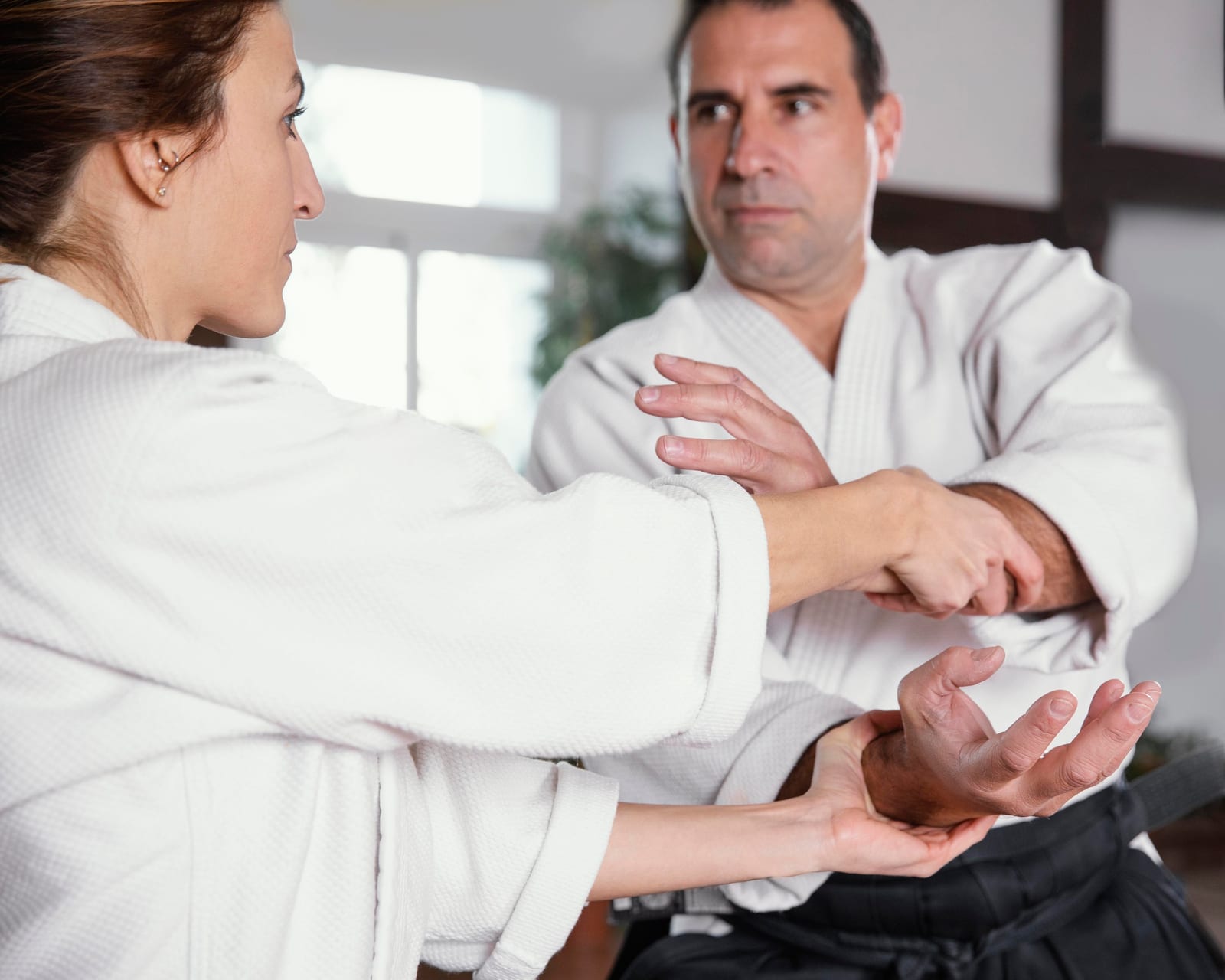 A woman in a white gi applies a wrist lock on a male instructor in a gi during a self-defense drill indoors.