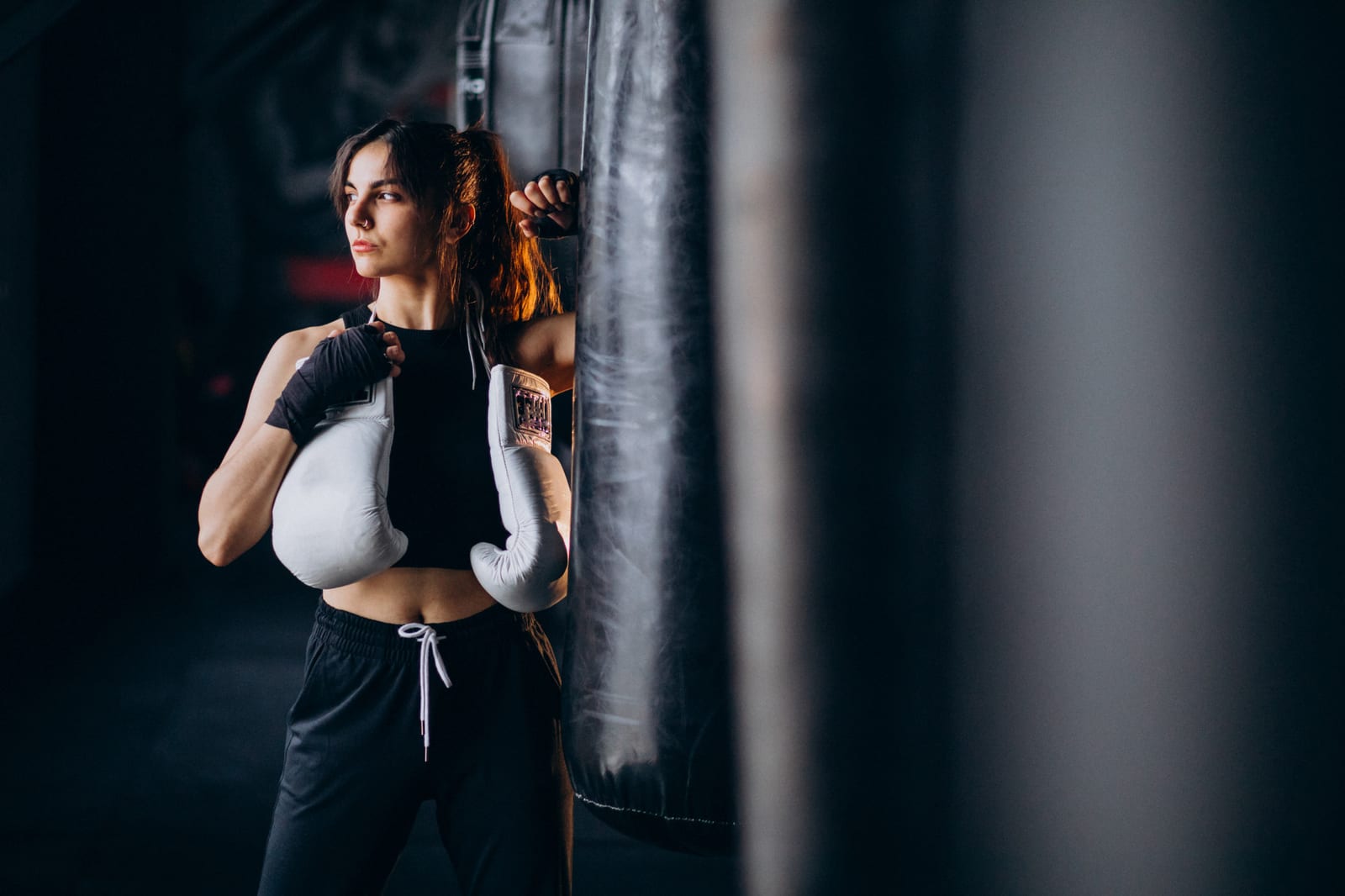A woman in boxing gloves and a black top stands in guard beside a heavy bag in a dark gym.