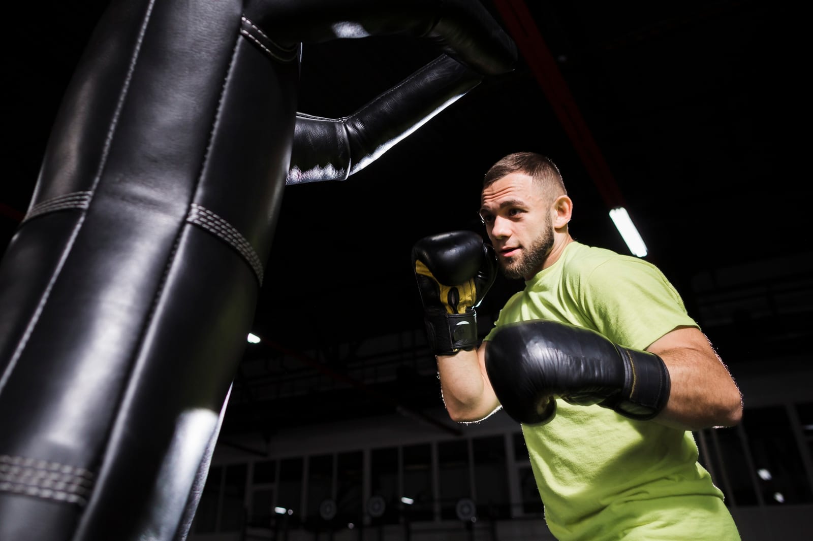 A man in a green shirt and boxing gloves throws a punch among hanging heavy bags in a dim gym.