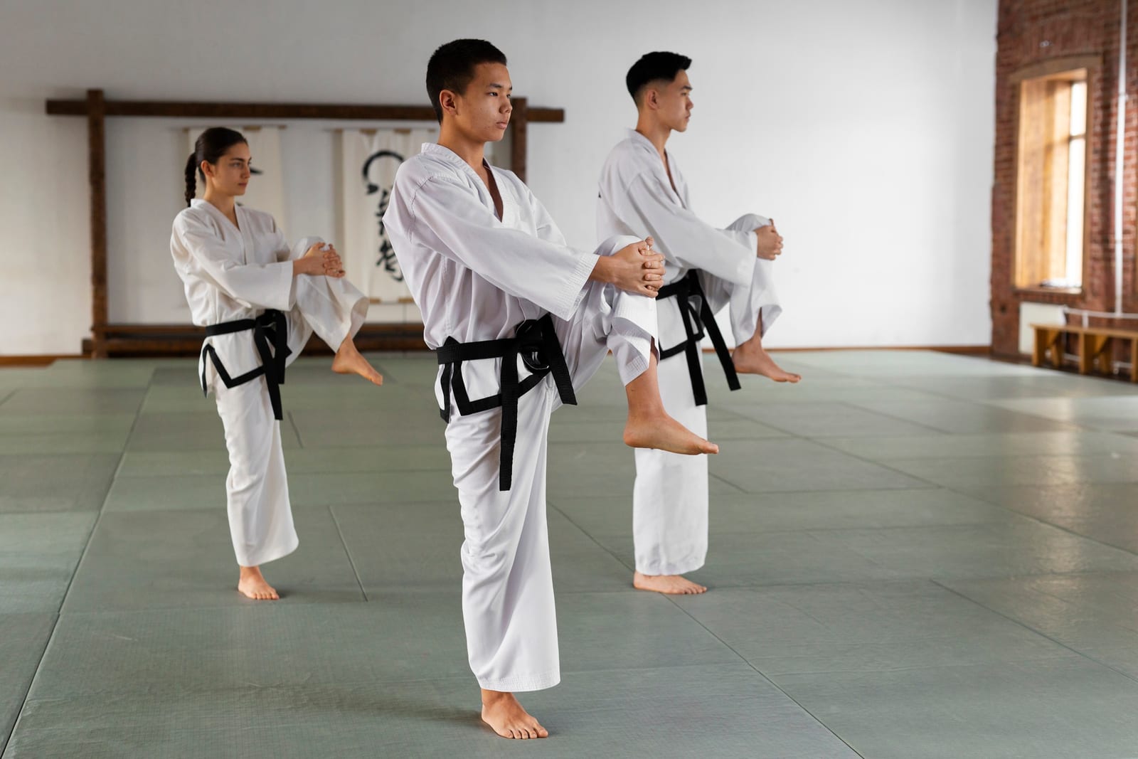 Three students in white taekwondo uniforms with black belts hold a raised knee position in a bright dojang.
