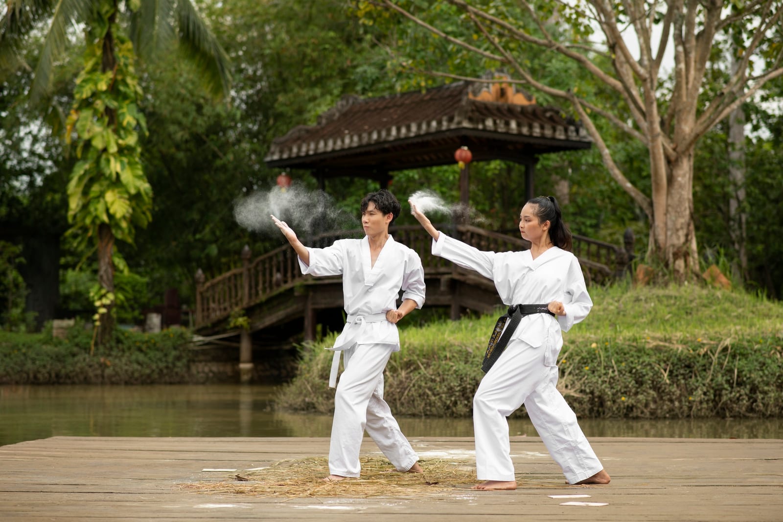 Two adults in white karate gis perform a kata on a wooden deck beside a pond outdoors.