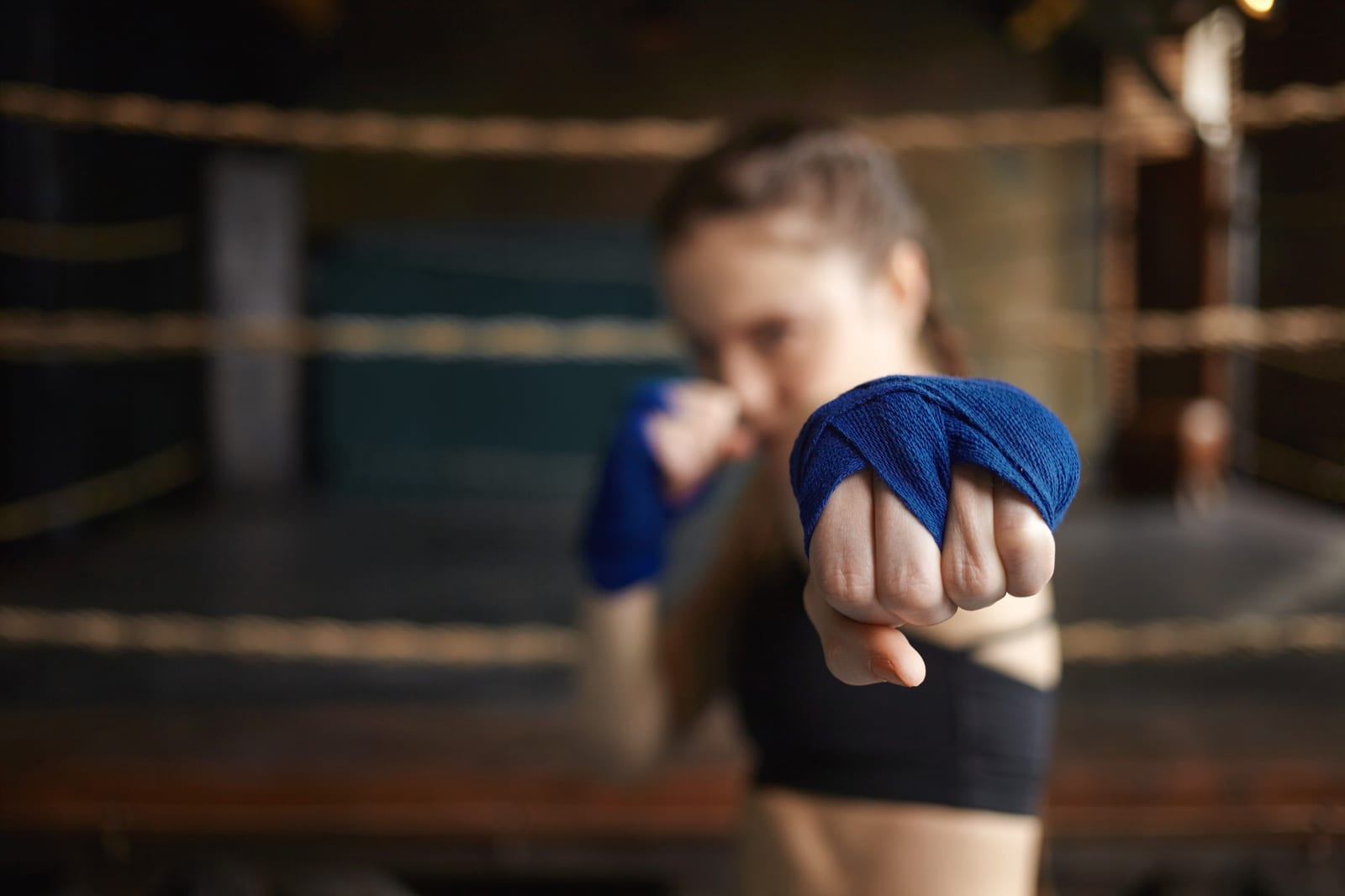 A woman with blue hand wraps holds a boxing guard inside a ring, the background softly blurred.