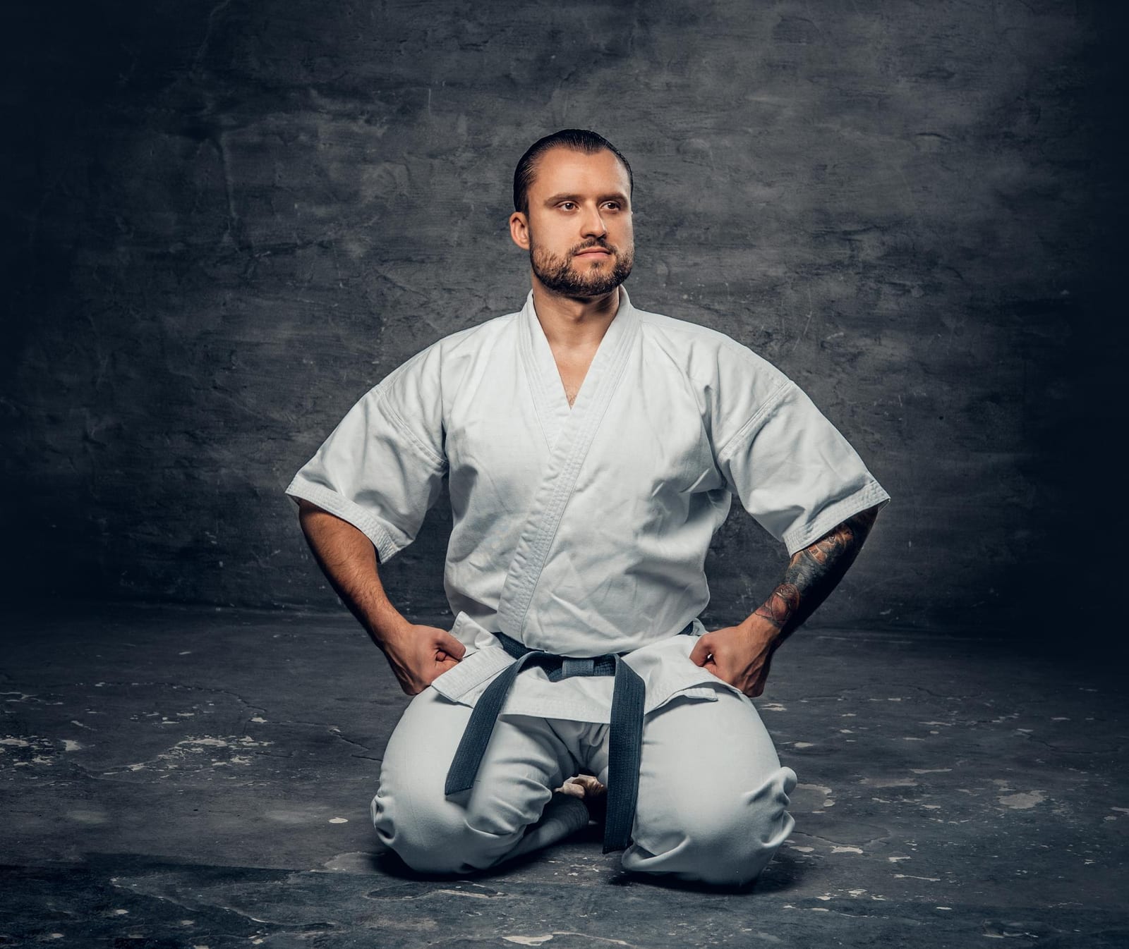 A man in a white karate gi with a grey belt kneels in seiza against a dark textured backdrop.